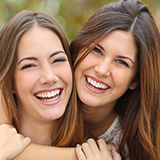 Two young brunette women with long hair hug each other while looking at the camera and smiling