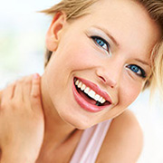 Blonde woman with short hair, blue eyes and big smile looks at the camera while having her hand on her neck