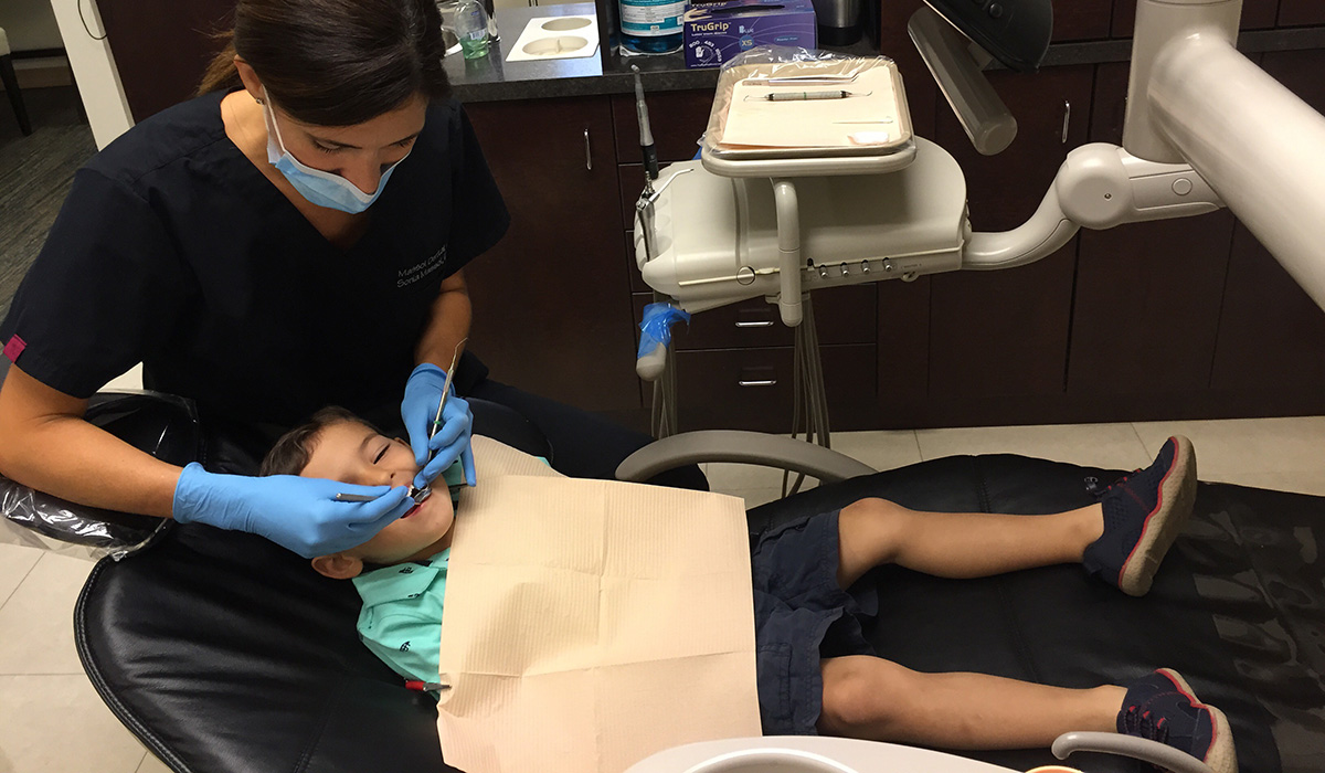 Dr. Sonia Massol in her dental office examines the mouth of a child who is wearing a green t-shirt and blue pants.
