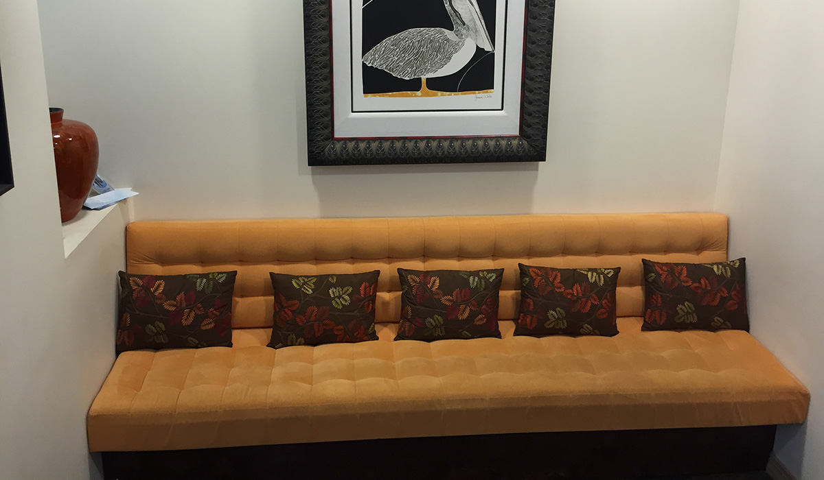 Waiting room in the office with a dark yellow sofa and five brown cushions with embroidered leaves and on the wall you can see half a painting of a white pelican on a black background