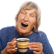 Senior woman with short hair and surprised face smiles at the camera while holding a cup of coffee in her hands