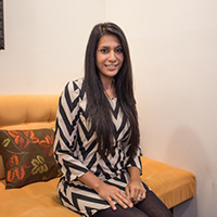 Woman with long black hair, sitting on a dark yellow sofa, wearing a dress with black and beige zig-zag lines, with brown pants